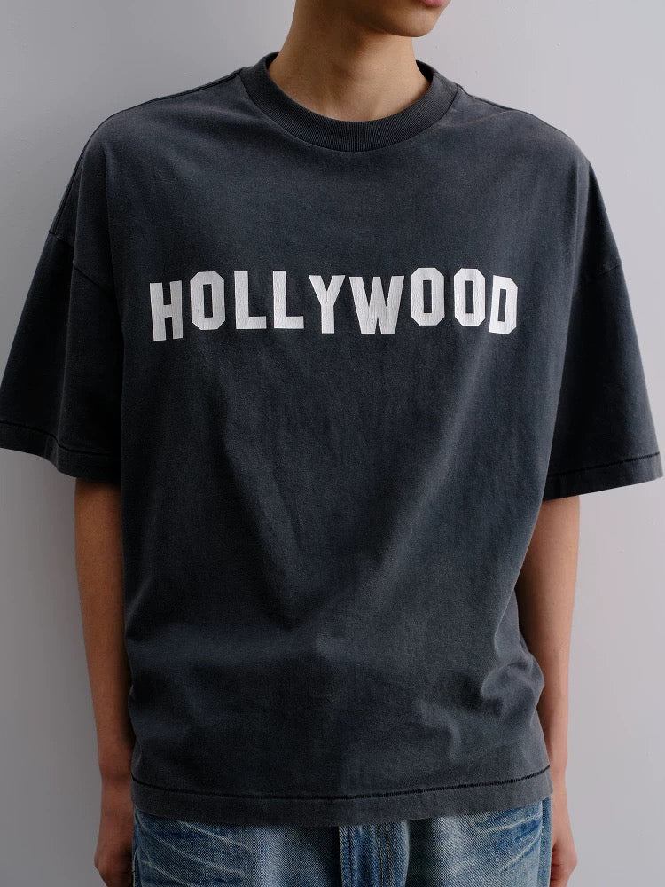 Person wearing a black t-shirt with 'HOLLYWOOD' printed on it against a gray background