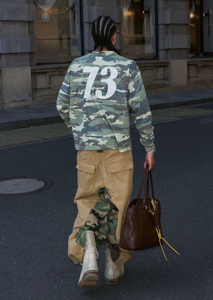 Person wearing a camouflage jacket with the number 73, walking on a street.