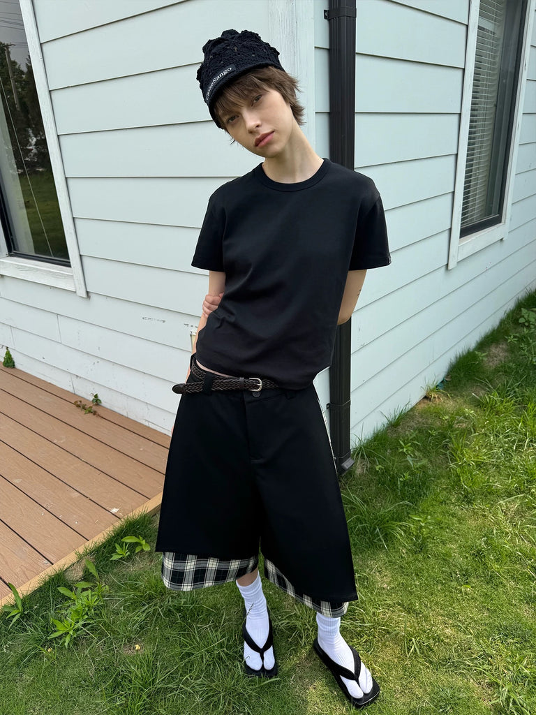 Person wearing a black outfit with plaid shorts standing outside a house.